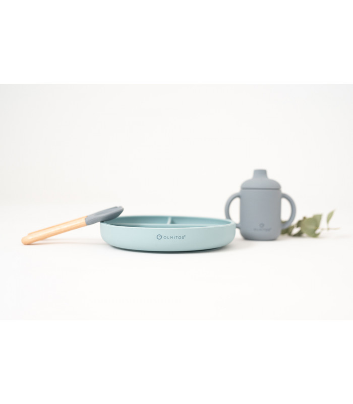 Vajilla de silicona Olmitos azul pastel con plato dividido, cuchara de madera y vaso con asas sobre un fondo blanco neutro.