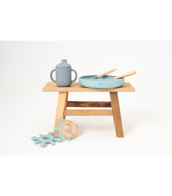 Set de 5 piezas de silicona azul Olmitos: plato con divisiones, taza, cubiertos y babero sobre un pequeño banco de madera.