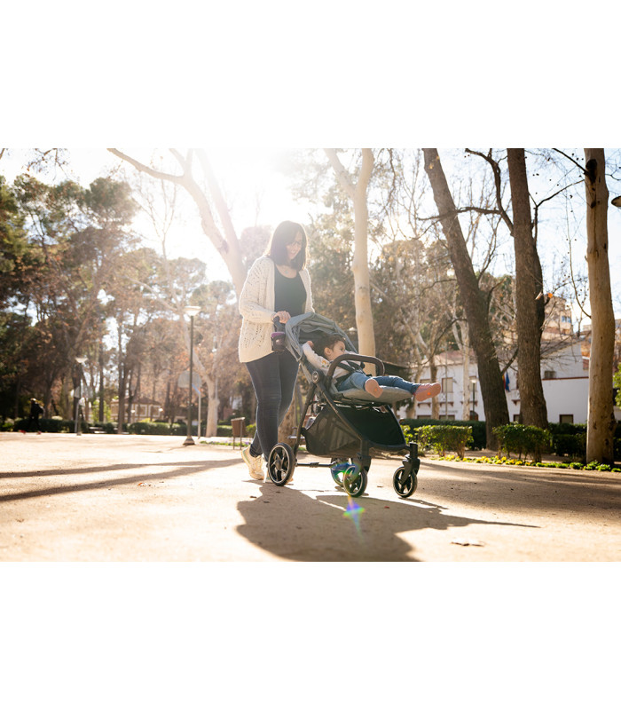 Silla de paseo VentT+ Niu negra con chasis metálico y capota, paseando por un parque soleado rodeado de árboles.