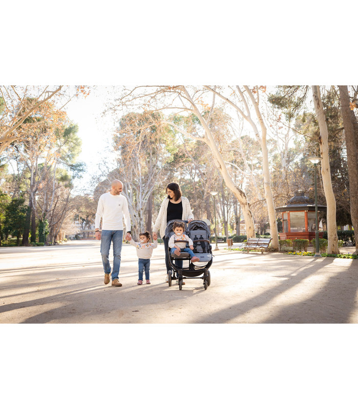 Silla gemelar VentTwin + Niu negra de estructura ligera, paseada por una familia en un parque arbolado y soleado.