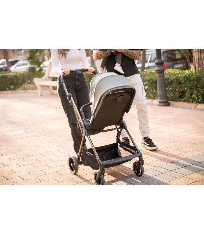 Silla de paseo Eolia Niu con chasis negro, capota gris claro y cesta inferior, paseando por un camino exterior soleado.