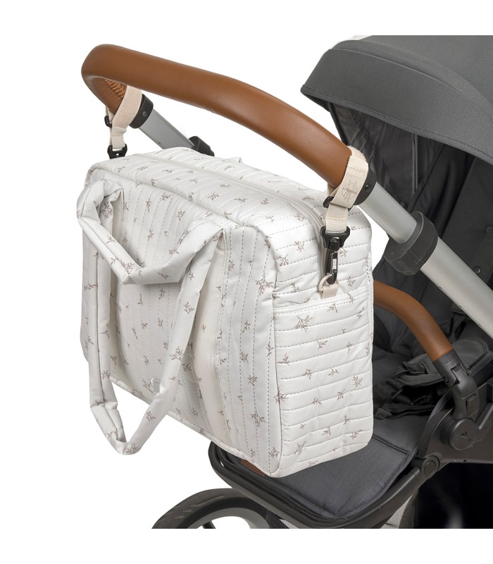 Bolso Nalea de Lässig en tela blanca con estampado sutil, colgado del manillar de cuero marrón de un cochecito de bebé.