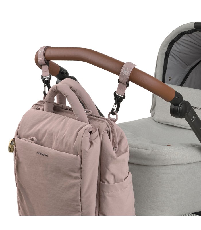 Mochila maternal Minoa de tela rosa palo, colgada mediante ganchos del manillar marrón de un cochecito gris.